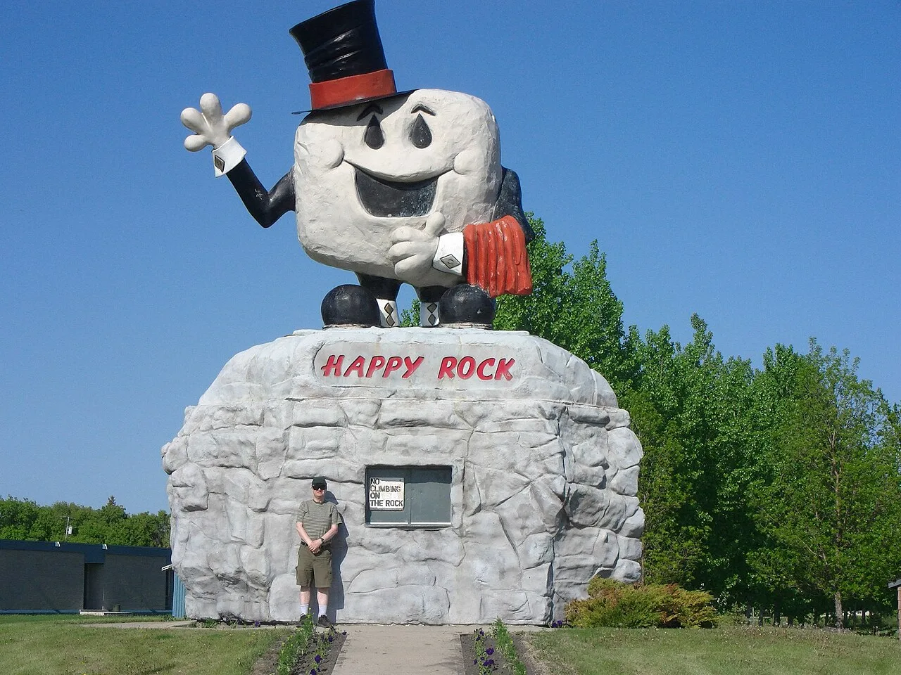 Photograph of Happy Rock in Gladstone, Manitoba, a grinning rock character in a top hat waving from the top of a large rock pedestal, with a visitor standing at its base