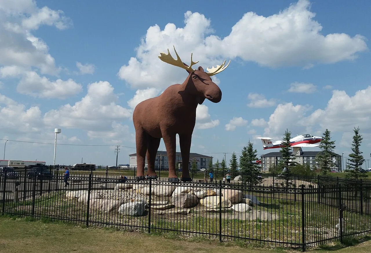 Photograph of Mac the Moose, the giant moose statue in Moose Jaw, Saskatchewan, standing on a rocky base inside a black iron fence under a blue sky